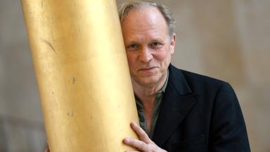 Ein Mann mit grauem Haar lehnt sich an einen goldenen Pfosten