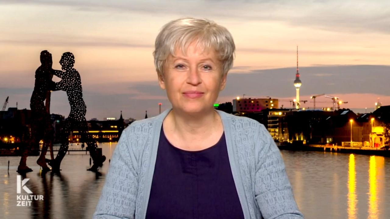 Eine Frau mit kurzen grau-blonden Haaren sitzt vor einer Berlin-Kulisse.