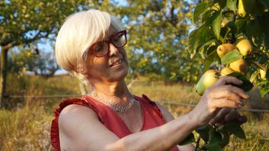 Eine Frau mit Brille pflückt lächelnd Birnen. 