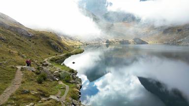 Eine malerische Berglandschaft im Piemont mit einem klaren See, dessen Wasser die umliegenden Berge und den Himmel mit Wolken spiegelt. Ein schmaler Wanderweg schlängelt sich am Ufer entlang, auf dem Wanderer zu sehen sind.