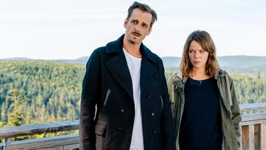 Ein Mann und eine Frau stehen vor einem Holzgeländer mit Wald im Hintergrund.