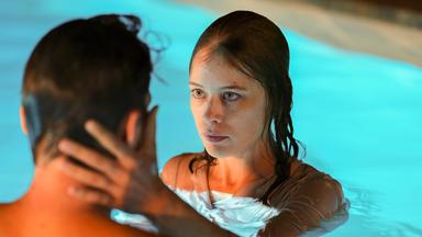 Junge Frau ist mit jungem Mann in einem Pool. Sie hält ihre linke Hand an seinen Kopf und schaut ihm in die Augen.