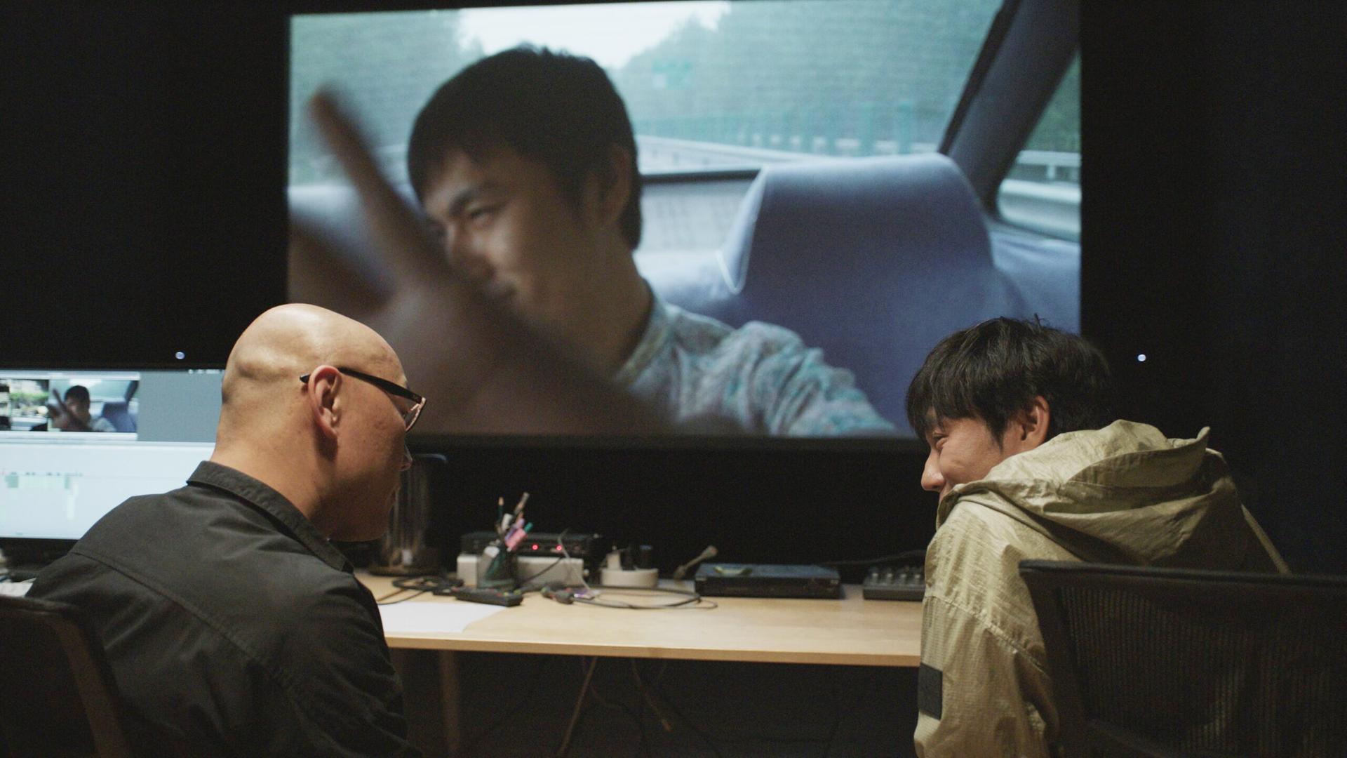 Zwei Männer sitzen vor einem Bildschirm, auf dem ein Mann in einem Auto zu sehen ist.