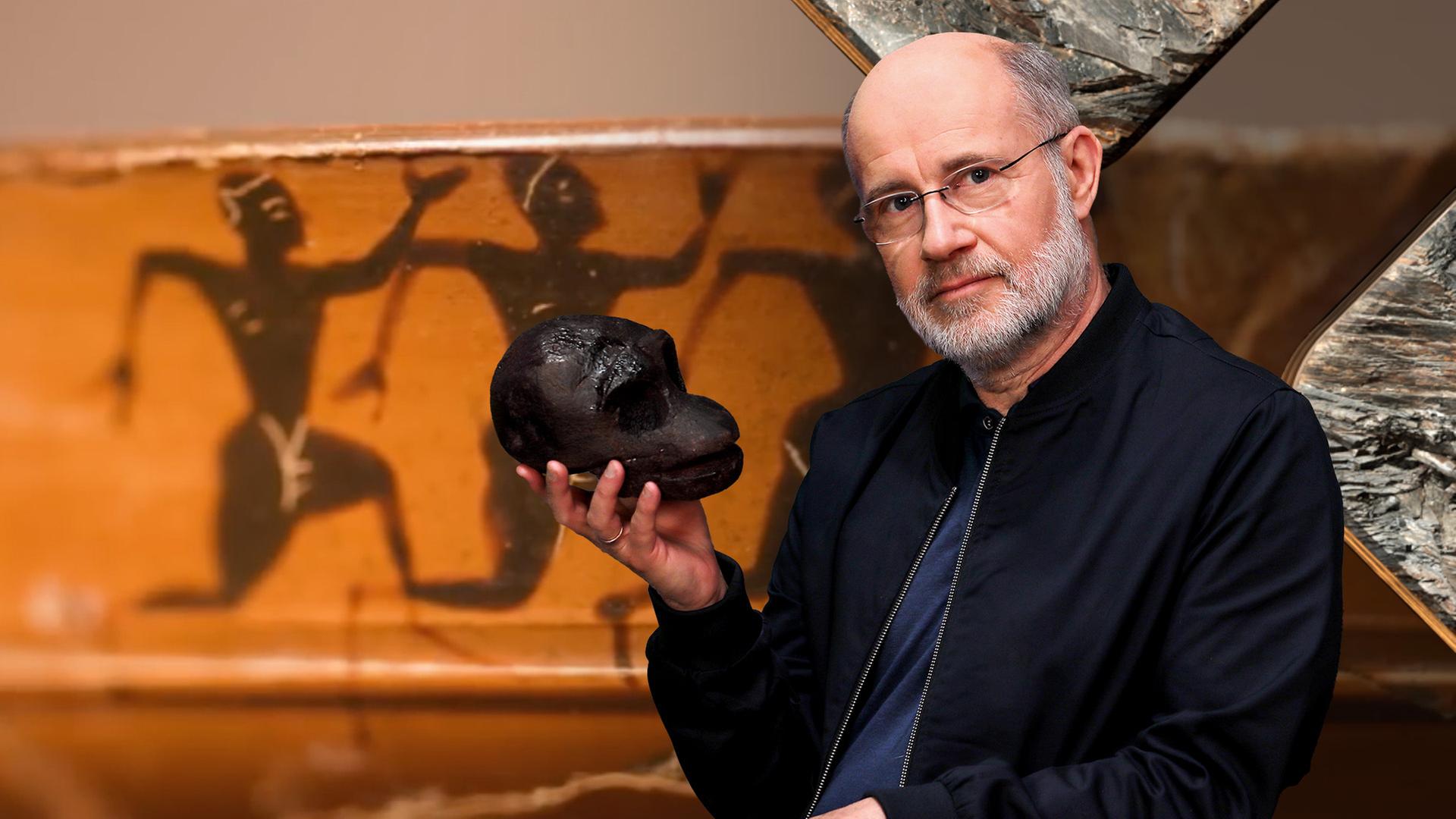 Harald Lesch hält eine schwarze Skulptur eines Schädels in der Hand. Hinter ihm ist eine Wandmalerei zu sehen, die eine Reihe von Frauen in Bewegung zeigt.