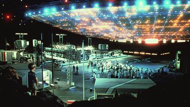 „Unheimliche Begegnung der dritten Art“: Ein riesiges, hell leuchtendes Raumschiff steht knapp über dem Boden, unter ihm steht eine größere Gruppe von Wissenschaftlern in weißer Kleidung, umringt von technischen Aufbauten. Auf der linken Bildseite ist ein kleiner Junge mit seiner Mutter zu sehen, die sich das Spektakel anschauen.