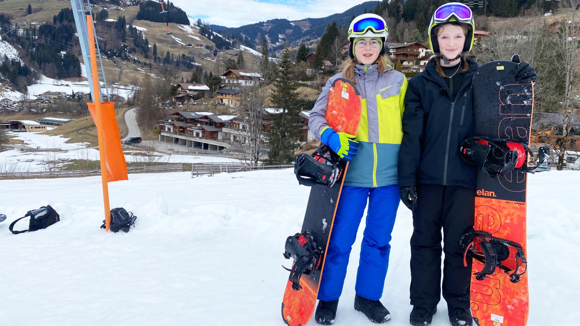 Zwei Mädchen stehen mit Snowboards auf einer Piste. Hinter ihnen eine Berglandschaft mit wenig Schnee.