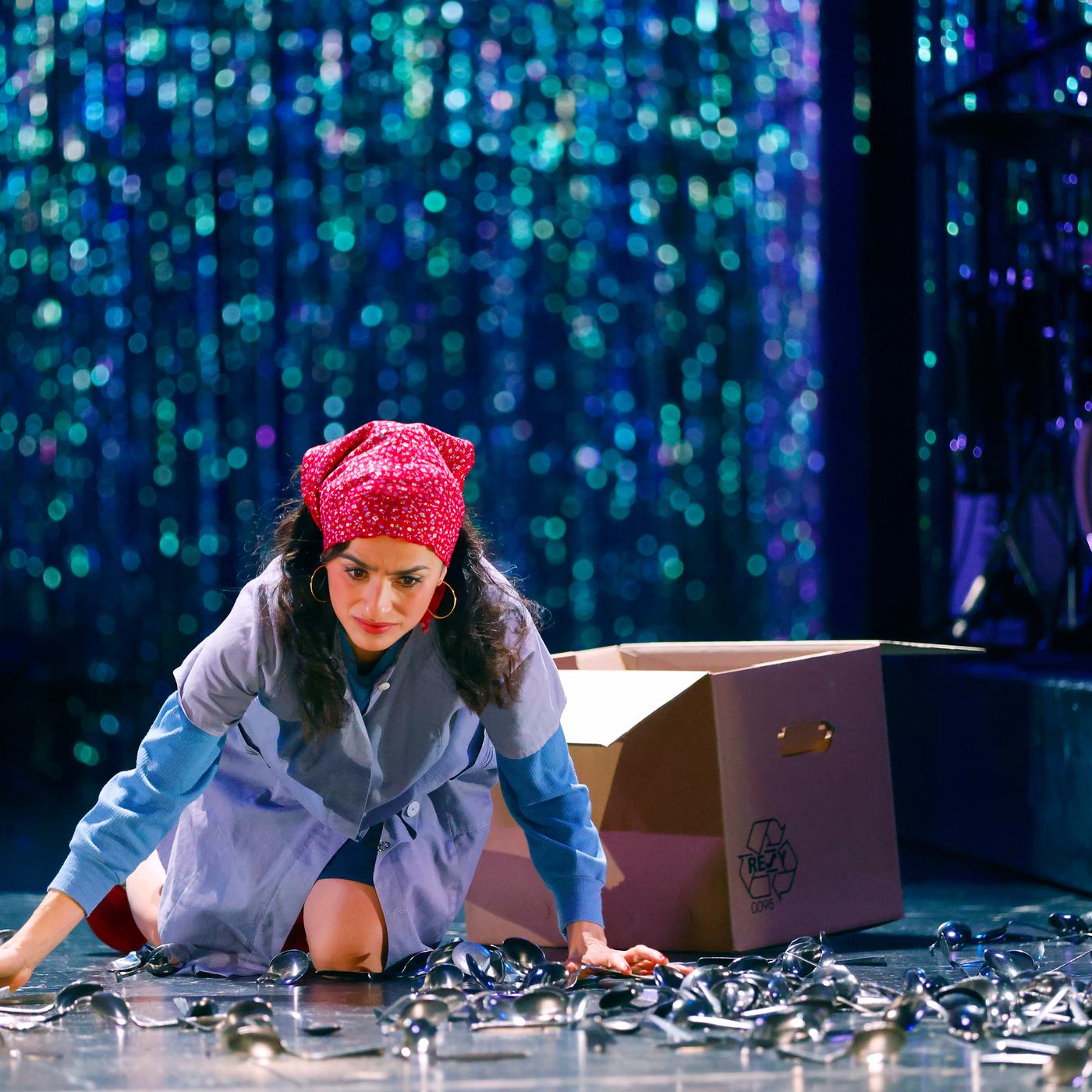 Schauspielerin in hellblauem Arbeitskittel, rotem Kopftuch mit weißen Punkten und blauen Ärmeln kniet auf einer Bühne, umgeben von verstreuten Löffeln. Sie stützt sich mit den Händen ab und blickt besorgt nach unten. Hinter ihr steht ein geöffneter Pappkarton mit Recycling-Symbol. Im Hintergrund ist ein glitzernder Bühnenvorhang zu erkennen, in der rechten oberen Eckw sind Teile eines Schlagzeugs und eines Musikers zu sehen.