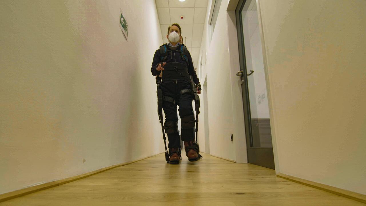 Das Bild zeigt eine Person, die in einem schmalen, hellen Flur geht. Die Wände sind weiß und der Boden ist aus hellem Holz. Die Person trägt eine Maske und hat einen Rucksack auf. Sie ist in einem speziellen Anzug, der Unterstützung beim Gehen bietet. Der Flur wird von einer Glastür an einer Seite und einem Notausgangsschild an der Wand entlang der anderen Seite gesäumt. Licht tritt von der Decke ein und schafft eine helle, klinische Atmosphäre.