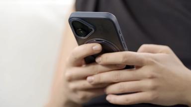 Das Bild zeigt Hände, die ein Smartphone halten. Die Person trägt ein schwarzes Oberteil. Der Hintergrund ist unscharf und hell. Das Smartphone hat eine ausgeprägte Schutzhülle.
