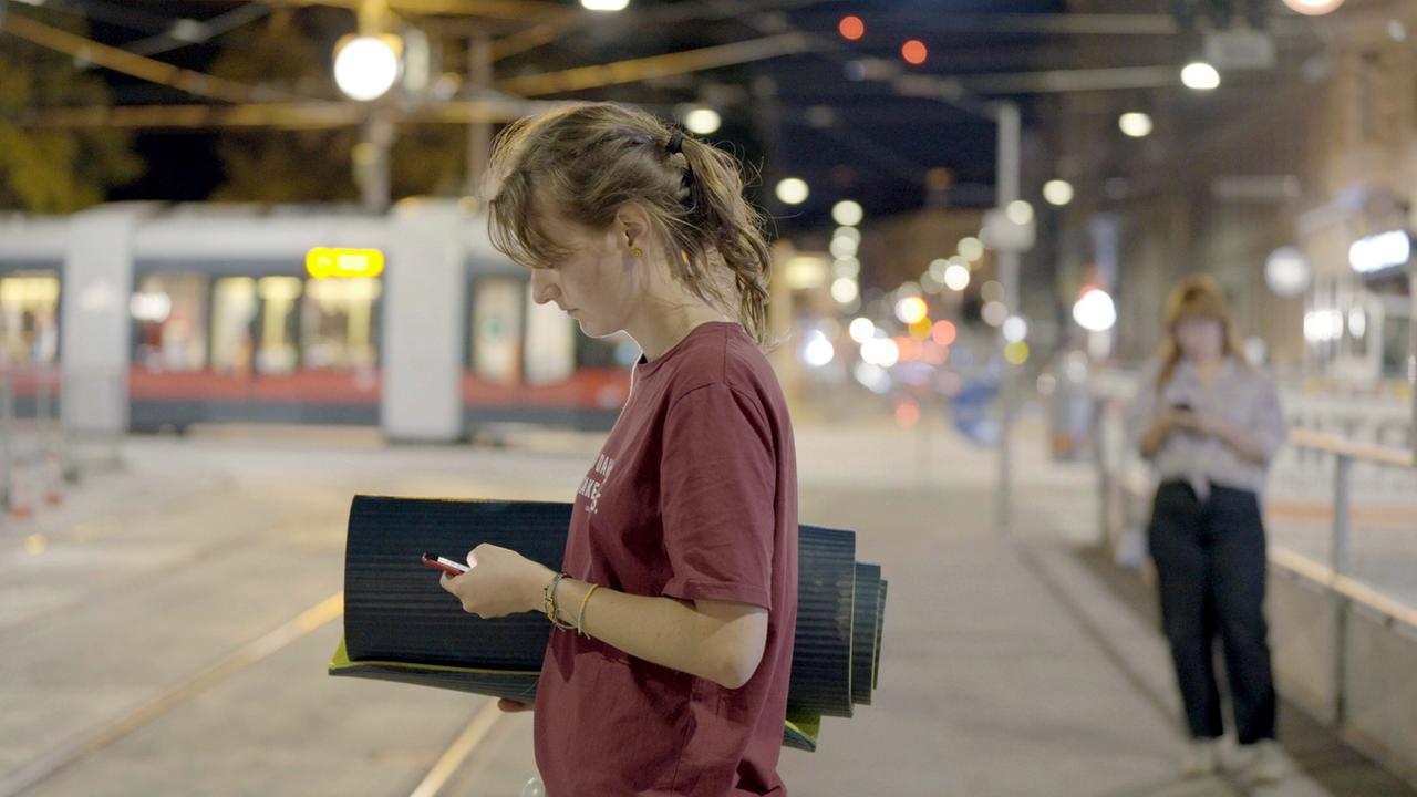 Eine Studentin steht an einer Straßenbahnhaltestelle, hält mehrere schwarze Matten und schaut auf ihr Handy. Im Hintergrund sind unscharf weitere Personen und Lichter zu sehen.