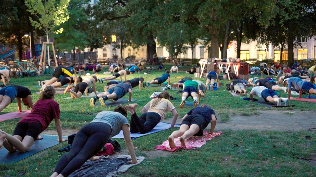 Eine Gruppe von Menschen macht Übungen auf Fitnessmatten im Freien. Der Platz ist von Bäumen umgeben, und einige Gebäude sind im Hintergrund sichtbar. Es ist Abendlicht.