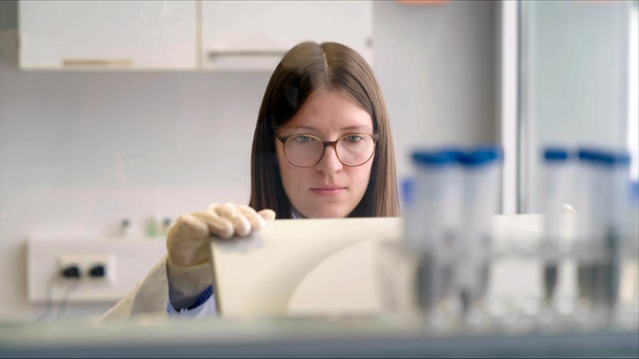 Eine Person in einem Labor schaut konzentriert auf ein Objekt, während sie Handschuhe trägt. Im Hintergrund sind mehrere Reagenzgläser und Laborutensilien zu sehen.