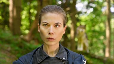Das Bild zeigt eine Frau mit kurzen, braunen Haaren und einem ernsten Gesichtsausdruck. Sie trägt eine schwarze, ledere Jacke. Der Hintergrund ist ein Wald mit grünen Bäumen und verschwommenen Lichtstrahlen. Hinter der Frau ist schemenhaft ein Wolf zu sehen, der ebenfalls im Wald steht. Das Bild vermittelt eine spannungsgeladene Atmosphäre, passend zu einem Kriminalfall, der in der Natur spielt.