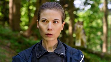 Das Bild zeigt eine Frau mit kurzen, braunen Haaren und einem ernsten Gesichtsausdruck. Sie trägt eine schwarze, ledere Jacke. Der Hintergrund ist ein Wald mit grünen Bäumen und verschwommenen Lichtstrahlen. Hinter der Frau ist schemenhaft ein Wolf zu sehen, der ebenfalls im Wald steht. Das Bild vermittelt eine spannungsgeladene Atmosphäre, passend zu einem Kriminalfall, der in der Natur spielt.