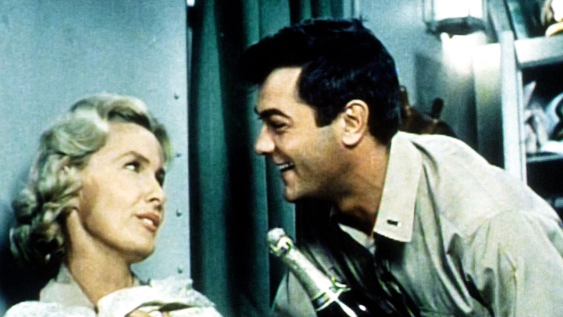 Ein Mann und eine Frau in der Kabine eines Schiffs. Er hält eine Sektflasche in der Hand.