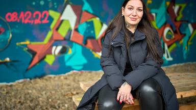 Streetworkerin Amelie Sophie Preyss sitzt auf einem Holztisch vor einer Graffiti-Wand. Sie trägt schwarze Kleidung, Tattoos an Hals und Händen, Piercings und hat rote Fingernägel.