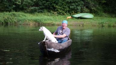 Ein Mann mit einer bunten Kopfbedeckung sitzt zusammen mit einem weißen Hund in einem Einbaumboot auf einem Fluss, umgeben von üppigem Grün.