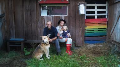 Eine Familie mit einem Hund sitzt vor einem Holzhaus. Die Frau hält ein kleines Kind auf dem Schoß. Neben der bunt gestrichenen Tür hängt eine Milchkanne.