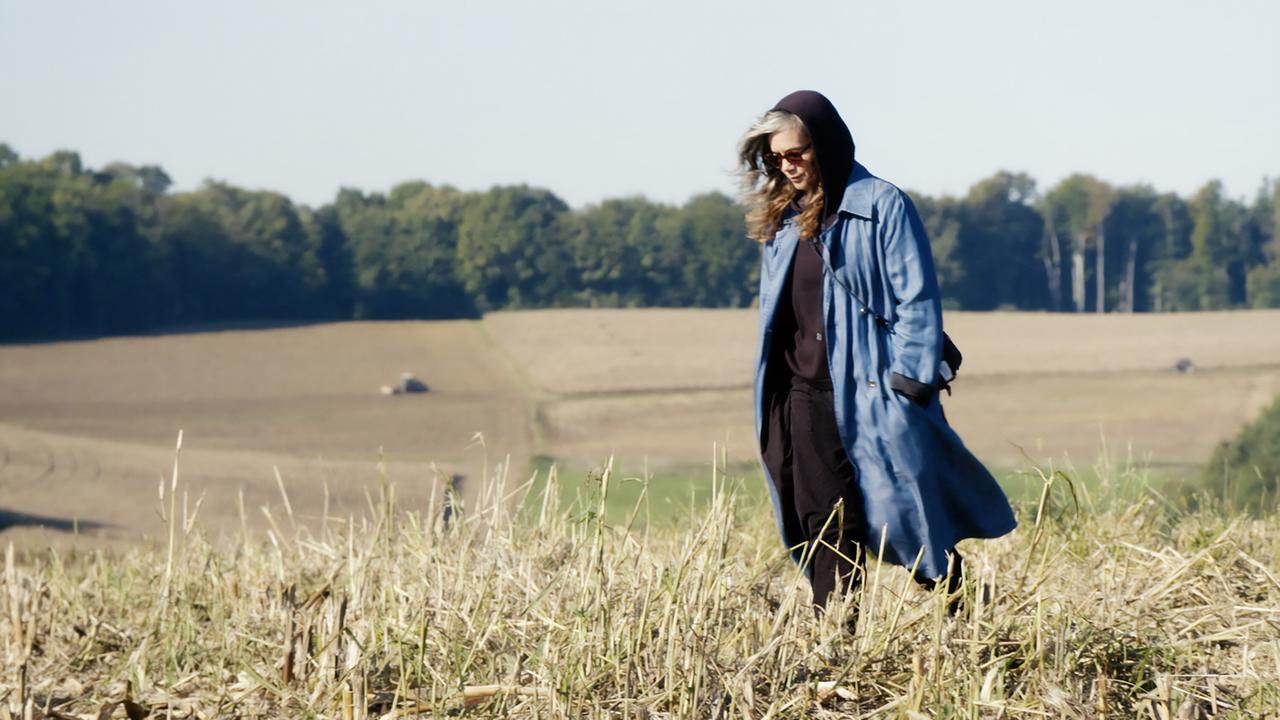 Das Bild zeigt eine Person, die auf einem abgeernteten Feld spaziert. Die Landschaft ist von sanften Hügeln und Bäumen umgeben, die im Hintergrund sichtbar sind. Der Himmel ist klar und es scheint sonnig zu sein. Die Person trägt eine lange, blaue Jacke mit einem Kapuzenpulli darunter und hat dunkle Hosen an. Ihr Haar ist lang und lockig, und sie trägt eine Sonnenbrille. Die Umgebung ist ruhig und ländlich, mit Streifen aus erhobenem Ackerboden und einigen Bäumen im Hintergrund.