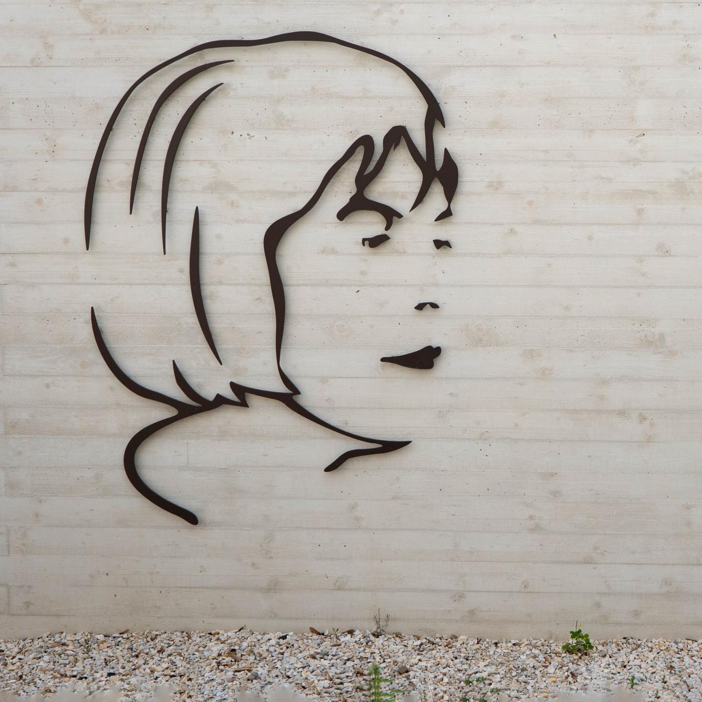Das Bild zeigt eine Wand des Ingeborg Bachmannparks, die aus hellem Beton besteht. Darauf ist ein stilisiertes, schwarzes Relief einer Gesichtsprofilansicht, das an Ingeborg Bachmann erinnert. Rechts neben dem Relief befindet sich ein schwarzer Tragbeutel, der mit einer grauen Zeichnung eines Gesichts und roten Texten geschmückt ist. Darunter steht die Zahl "50". Über dem Relief und der Tasche ist das Logo „ORF K“ in einem schlichten, modernen Design angebracht. Der Boden vor der Wand besteht aus kleinen, hellen Steinen und es sind einige grüne Pflanzen zu erkennen.