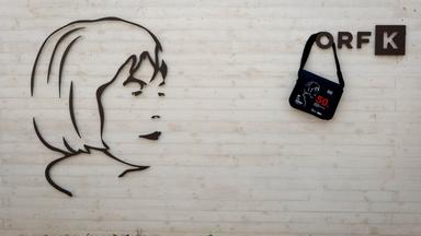 Das Bild zeigt eine Wand des Ingeborg Bachmannparks, die aus hellem Beton besteht. Darauf ist ein stilisiertes, schwarzes Relief einer Gesichtsprofilansicht, das an Ingeborg Bachmann erinnert. Rechts neben dem Relief befindet sich ein schwarzer Tragbeutel, der mit einer grauen Zeichnung eines Gesichts und roten Texten geschmückt ist. Darunter steht die Zahl "50". Über dem Relief und der Tasche ist das Logo „ORF K“ in einem schlichten, modernen Design angebracht. Der Boden vor der Wand besteht aus kleinen, hellen Steinen und es sind einige grüne Pflanzen zu erkennen.