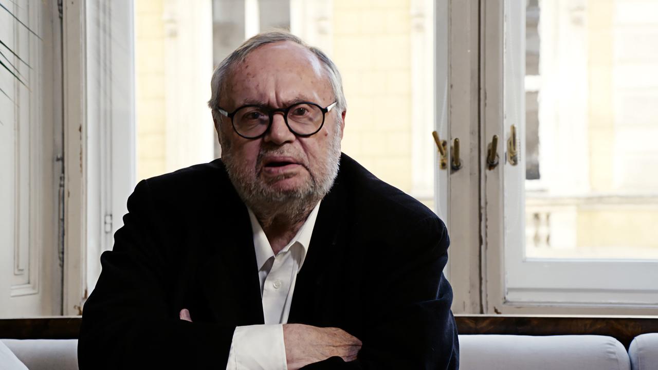 Das Bild zeigt einen älteren Mann mit Brille, der in einem Raum sitzt. Er trägt einen schwarzen Blazer über einem weißen Hemd. Der Mann hat graue, leicht zerzauste Haare und einen Bart. Er blickt direkt in die Kamera und hat die Arme vor sich verschränkt. Hinter ihm sind große Fenster mit hellen Fensterrahmen zu sehen, durch die natürliches Licht scheint. Das Licht erhellt den Raum und wirft sanfte Schatten. Im Hintergrund sind unscharf gespannte, hellgelbe Wände zu erkennen. Der Gesamteindruck des Bildes vermittelt eine nachdenkliche, ernste Atmosphäre.