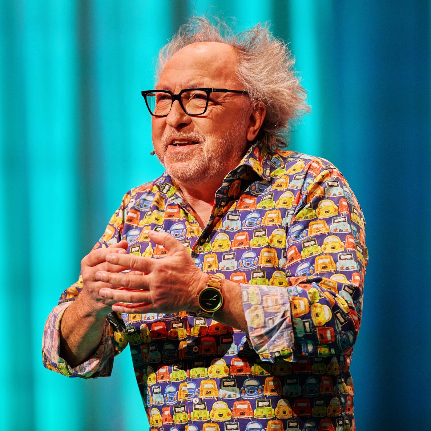 Urban Priol ist in der Mitte des Bildes zu sehen. Er hebt seine Hände vor seine Brust. Sein Hemd ist bunt. Urban Priol trägt eine Brille. Der Hintergrund ist türkis beleuchtet. Am linken Handgelenk trägt er eine Uhr.