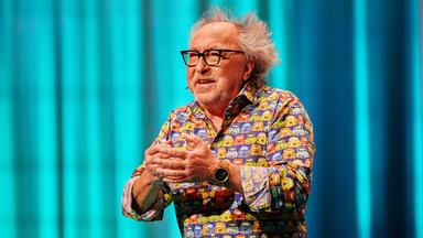 Urban Priol ist in der Mitte des Bildes zu sehen. Er hebt seine Hände vor seine Brust. Sein Hemd ist bunt. Urban Priol trägt eine Brille. Der Hintergrund ist türkis beleuchtet. Am linken Handgelenk trägt er eine Uhr.