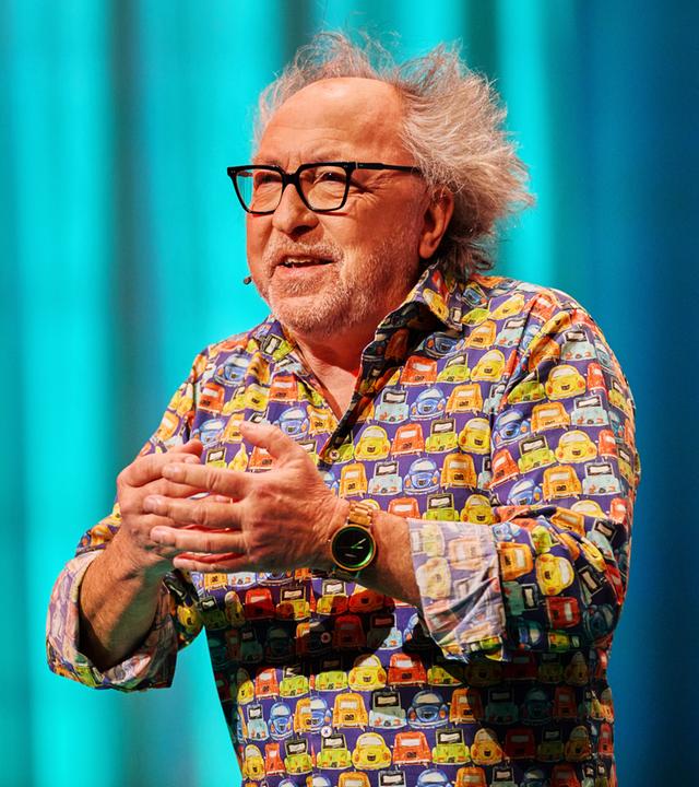 Urban Priol ist in der Mitte des Bildes zu sehen. Er hebt seine Hände vor seine Brust. Sein Hemd ist bunt. Urban Priol trägt eine Brille. Der Hintergrund ist türkis beleuchtet. Am linken Handgelenk trägt er eine Uhr.