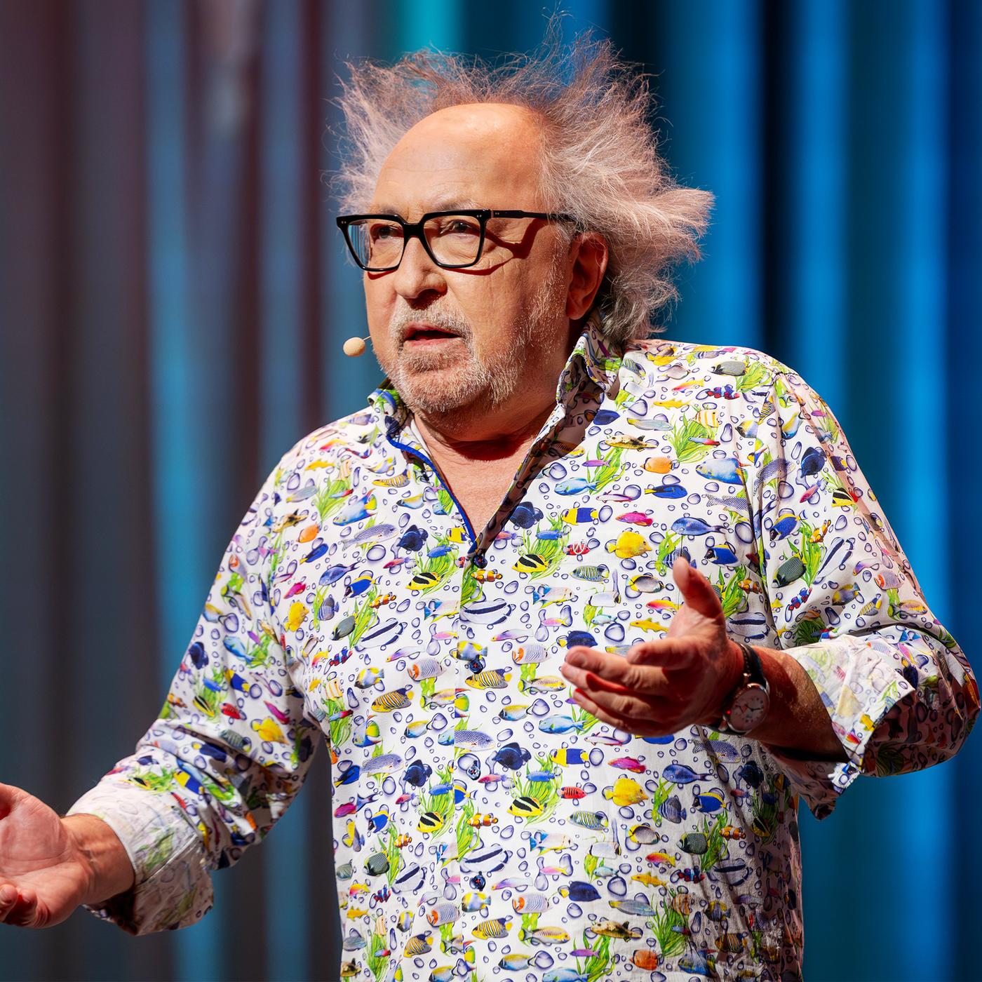 Urban Priol steht auf der Bühne. Er trägt ein buntes Hemd mit kleinen Fischen darauf. Er hält die Arme hoch. Der Hintergrund ist blau.