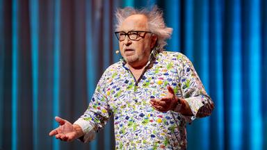 Urban Priol steht auf der Bühne. Er trägt ein buntes Hemd mit kleinen Fischen darauf. Er hält die Arme hoch. Der Hintergrund ist blau.