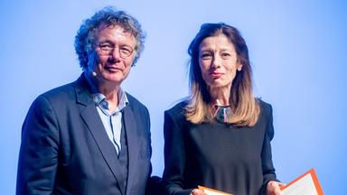 Eine Frau mit langen Haaren hält eine Kladde in den Händen, die sie geöffnet präsentiert. Neben ihr steht ein Mann.