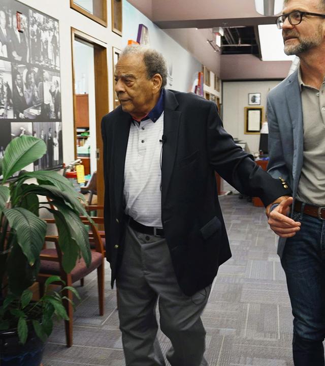 Peter Düggeli und Andrew Young gehen durch einen Flur, an der Wand sind historische Fotos zu sehen.