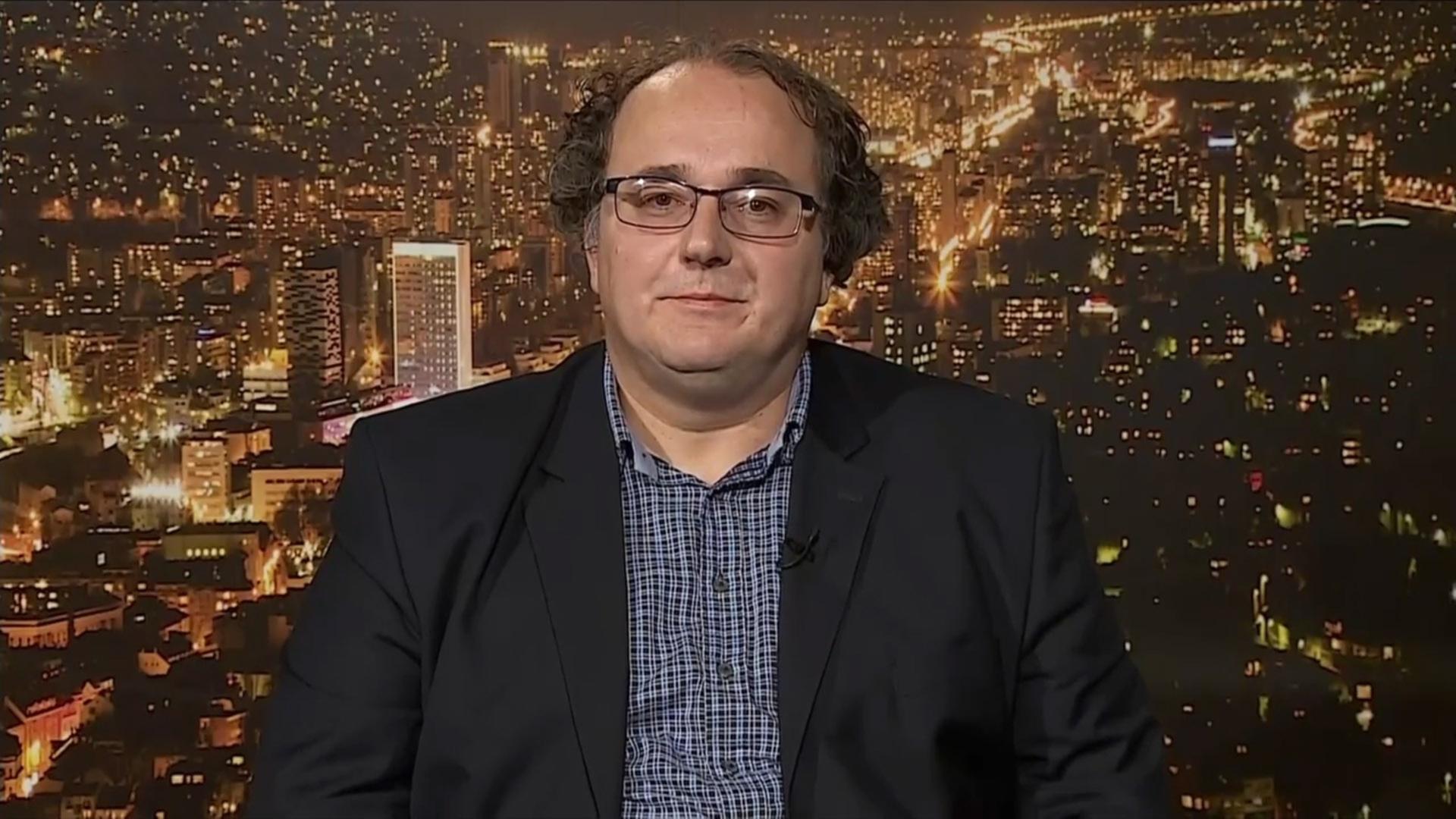 Ein Mann mit Brille in Anzug vor dem Hintergrund einer beleuchteten Stadt.