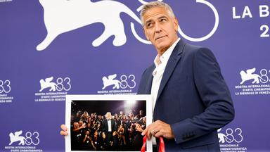Ein Mann im Anzug posiert vor der Fotowand des Filmfestivals Venedig mit einem Bild von sich selbst.