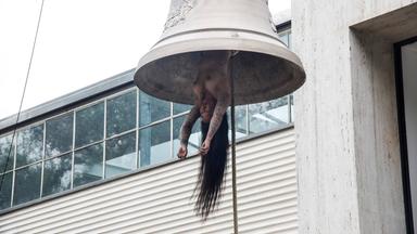 Eine nackte tätowierte Frau mit langen Haaren hängt wie ein Schlägel in einer Glocke.