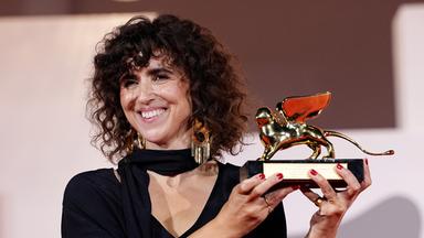 Eine Frau mit schulterlangen braunen Locken und im schwarzen Abendkleid präsentiert lächelnd eine goldene Trophäe, die einen geflügelten Löwen darstellt.