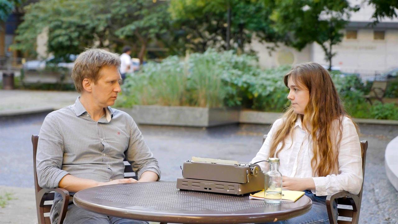 Auf dem Bild sitzen zwei Personen an einem runden Tisch im Freien. Vor ihnen steht eine alte Schreibmaschine. Beide sind in einem ruhigen Gespräch vertieft, umgeben von Grünpflanzen im Hintergrund.