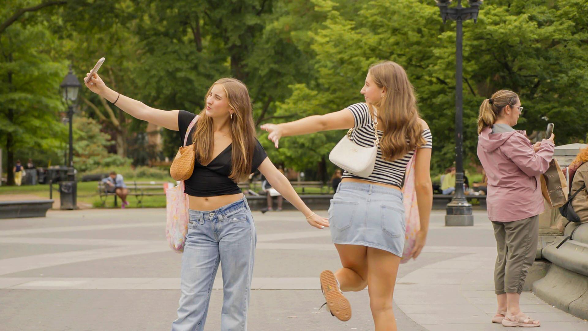 Zwei junge Frauen posieren im Washington Square Park in New York. Eine hält ein Smartphone in der Hand, während die andere eine Pose einnimmt. Im Hintergrund sind Bäume und Menschen zu sehen.