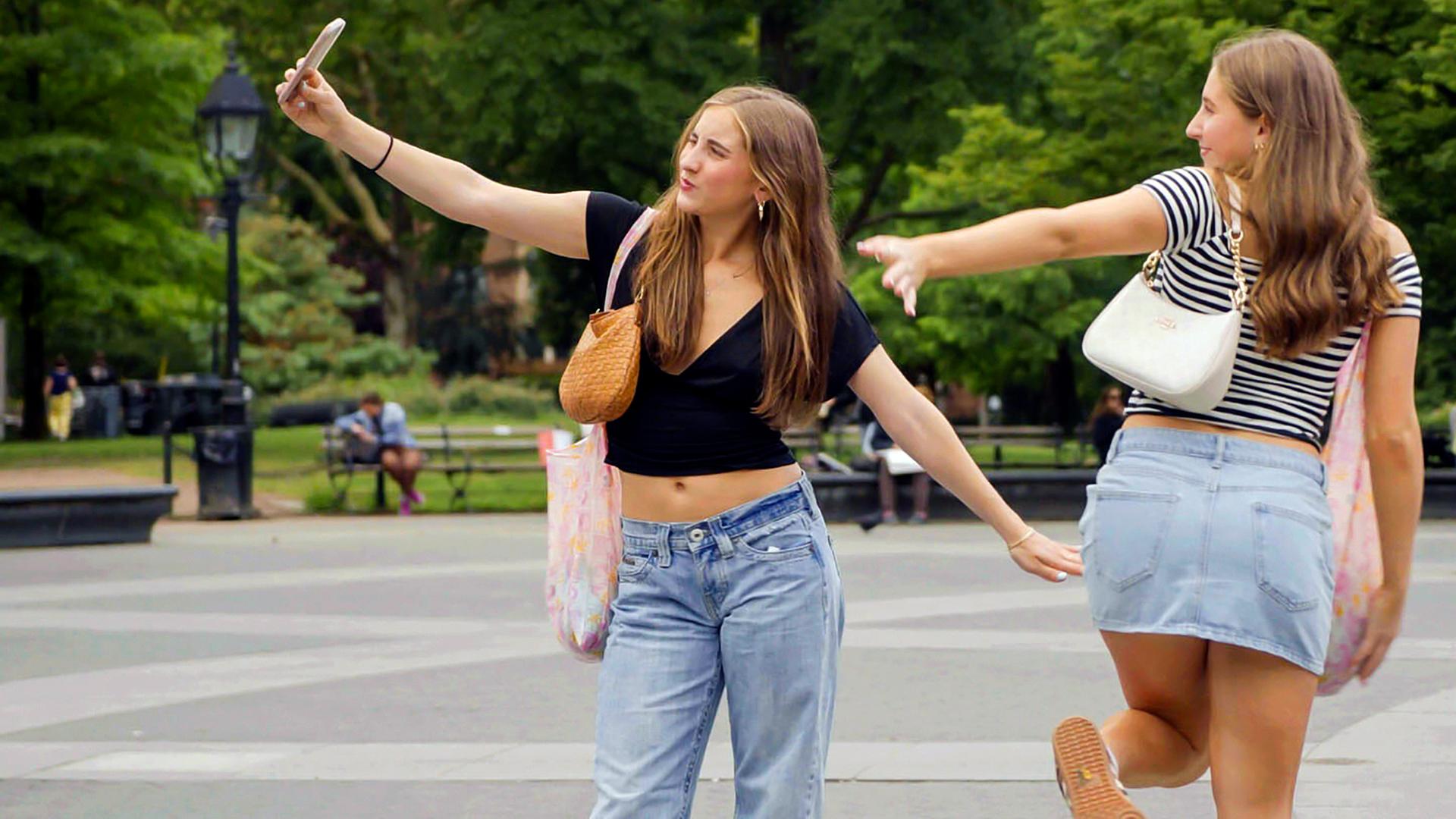 Zwei junge Frauen posieren im Washington Square Park in New York. Eine hält ein Smartphone in der Hand, während die andere eine Pose einnimmt. Im Hintergrund sind Bäume und Menschen zu sehen.