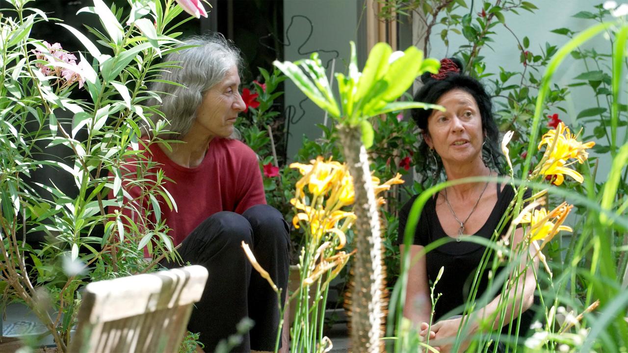 Das Bild zeigt zwei Frauen in einem privaten Biedermeiergarten. Im Vordergrund sind viele Pflanzen zu sehen, darunter gelbe Blumen, die in voller Blüte stehen. Die Frauen sitzen im Grünen, umgeben von verschiedenen Pflanzen und Sträuchern. Die eine Frau trägt ein rotes Oberteil, während die andere ein schwarzes Trägertop trägt. Beide scheinen entspannt und engagiert im Gespräch zu sein. Der Hintergrund ist weniger deutlich, da er mit Pflanzen und möglicherweise einer Wand oder einem Fenster verschwommen ist. Insgesamt vermittelt das Bild eine ruhige, naturnahe Atmosphäre inmitten eines gepflegten Gartens.