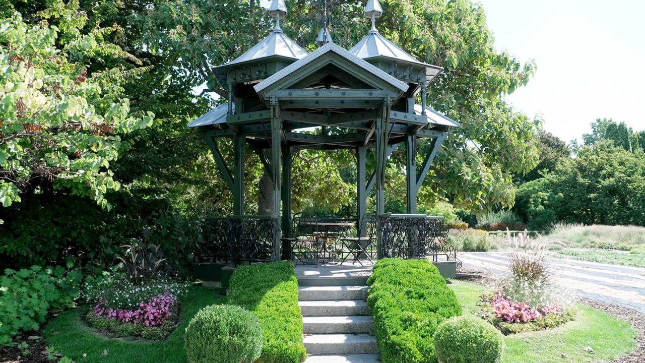 Das Bild zeigt einen Pavillon in einem Garten. Der Pavillon hat ein graues Dach und ist aus Holz mit verzierten Kanten und einem schwarzen Geländer gefertigt. Vor dem Pavillon führen ein paar Stufen zu einer ebenen Fläche. Um den Pavillon herum gibt es gepflegte Rasenflächen sowie einige Blumenbeete mit bunten Blumen, insbesondere rosa und weiße Blüten. Im Hintergrund sind hohe Bäume und Sträucher zu sehen, die dem Platz eine grüne und schattige Atmosphäre verleihen. Die Szene vermittelt einen idyllischen Eindruck eines ruhigen Gartens.