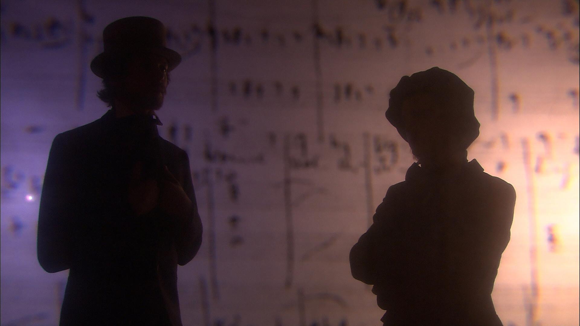 Das Bild zeigt zwei Personen in Silhouette, die vor einem verschwommenen, hellen Hintergrund stehen. Der Hintergrund könnte aus Notenblättern bestehen, da darauf Zeichen und Linien erkennbar sind. Die beiden Figuren tragen historische Kleidung aus dem 19. Jahrhundert: die linke Figur hat einen hohen Zylinderhut und eine dunkle Jacke, während die rechte Figur eine Kopfbedeckung und ein Kleidungsstück trägt, das an die damalige Mode erinnert. Die Beleuchtung des Bildes bewirkt, dass die Details der Personen schwer erkennbar sind, wodurch der Fokus auf der Interaktion zwischen den beiden und dem Hintergrund liegt.