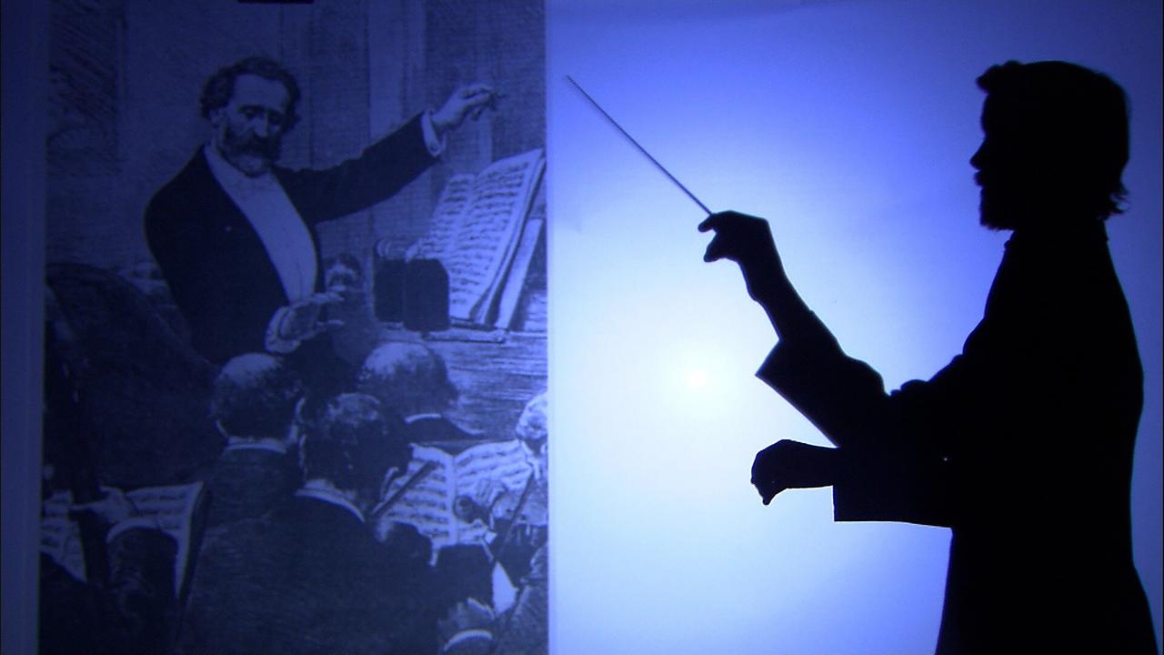 Das Bild zeigt eine künstlerische Darstellung, die zwei wesentliche Elemente enthält. Auf der linken Seite ist eine historische Schwarz-Weiß-Zeichnung zu sehen, die einen Dirigenten darstellt, der mit einem Taktstock dirigiert. Der Dirigent trägt einen Anzug und ist umgeben von einem Orchester, dessen Mitglieder von hinten zu sehen sind.  Rechts im Bild ist eine Silhouette einer Person, die ebenfalls mit einem Taktstock dirigiert. Diese Silhouette ist in dunklem Licht vor einem helleren, bläulichen Hintergrund abgebildet. Der Unterschied zwischen den beiden Darstellungen und die Kontraste in Licht und Schatten vermitteln eine dynamische Atmosphäre und regen zur Reflexion über die beiden schematisch dargestellten Dirigenten an.  Insgesamt vermittelt das Bild eine Verbindung zwischen Vergangenheit und Gegenwart in der Musik.