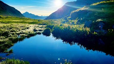 Ein kleiner See umgeben von Wildpflanzen, Steinen und Wiesen, eingebettet in hochaufragende Berge und Gipfel