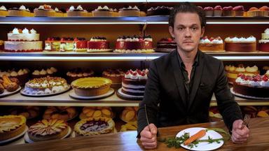 Jo Schück sitzt vor einem Teller mit einer Möhre, im Hintergrund ein Sortiment köstlicher Torten