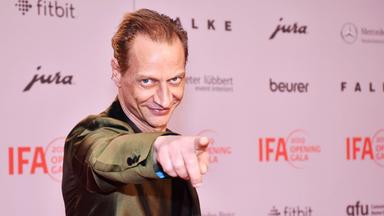 Foto von einem Mann im Anzug, der verschmitzt lächelnd mit dem Finger auf den Betrachter zeigt.