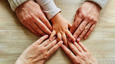 Fünf Hände kommen von unterschiedlichen Seiten ins Bild und berühren sich in der Mitte. Die Hände eines Mannes, einer Frau und eine Kinderhand.