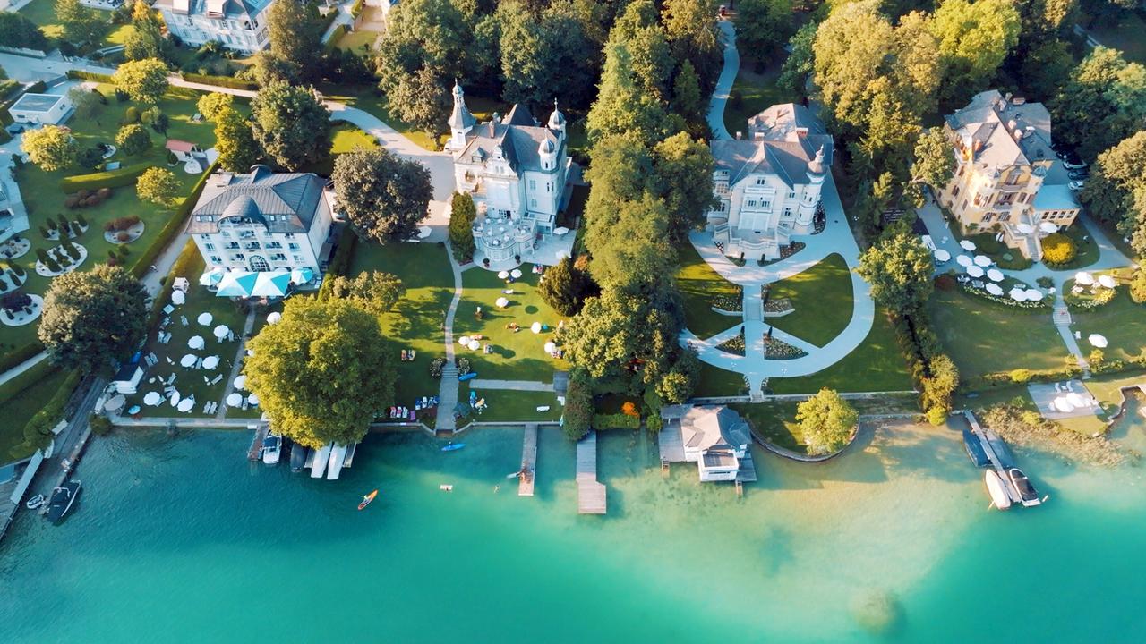 Das Bild zeigt eine Luftaufnahme des Wörthersees in Österreich, umgeben von prächtigen Villen. Die Villen sind architektonisch unterschiedlich, viele im sogenannten Wörthersee-Stil, der Elemente des Jugendstils und der Regionalromantik kombiniert.   Im Vordergrund erkennt man eine Strandpromenade mit Bootsanlegern aus Holz, die ins glitzernde, türkisfarbene Wasser führen. Einige Personen sind am Ufer verteilt, unter Schirmen oder auf Liegen und genießen die Umgebung. Im Hintergrund sind mehrere große Villen zu sehen, umgeben von begrünten Gärten und Bäumen. Dazu führen geschwungene Wege, die durch die Anlage verlaufen. Die gesamte Szene vermittelt eine entspannte, sommerliche Atmosphäre.