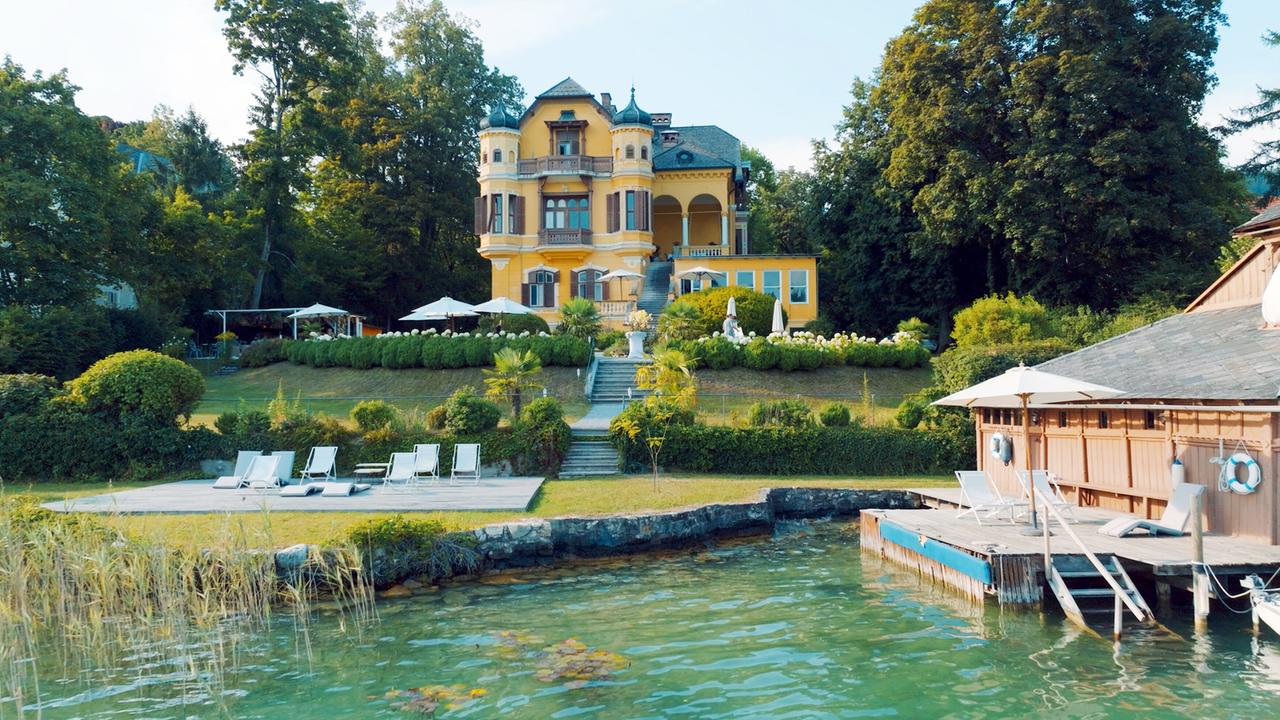 Das Bild zeigt die Villa Miralago am Wörthersee, umgeben von üppigem Grün. Die Villa ist mehrstöckig und hat eine gelbe Fassade mit mehreren großen Fenstern und Erkern. Auf dem Dach stehen zwei turmartige Elemente. Vor der Villa führt eine steile Treppe zu einem Garten mit gepflegten Beeten und verschiedenen Pflanzen. In der Nähe des Seeufers sind Liegen auf einem Holzdeck angeordnet, und gegenüber steht eine hölzerne Plattform mit Sonnenschirmen. Das Wasser des Wörthersees ist ruhig und hat eine grünliche Farbe. Die Umgebung vermittelt eine ruhige und elegante Atmosphäre.